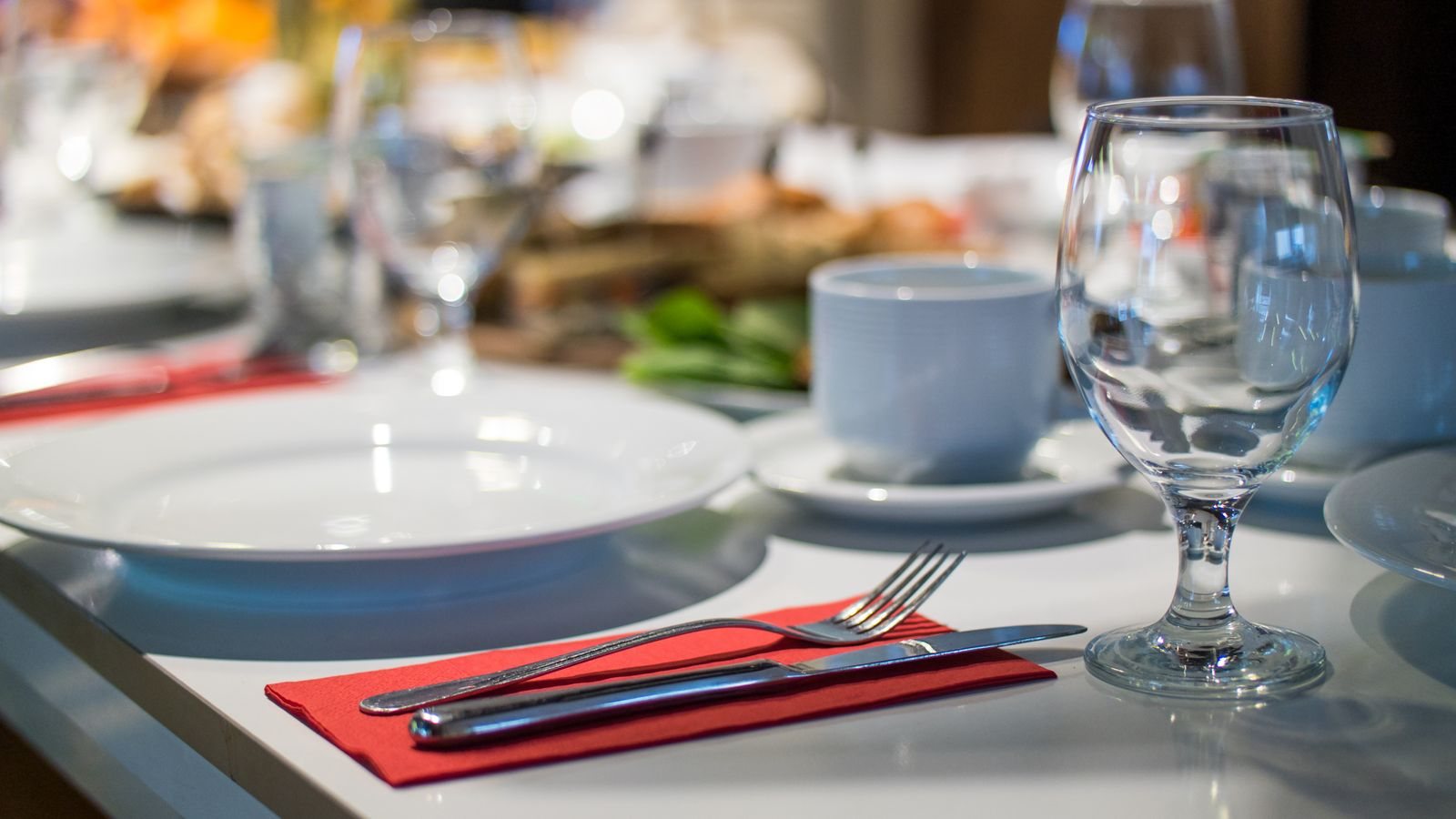 Masă de restaurant aranjată elegant, cu farfurii albe, pahar de vin și tacâmuri pe un șervet roșu