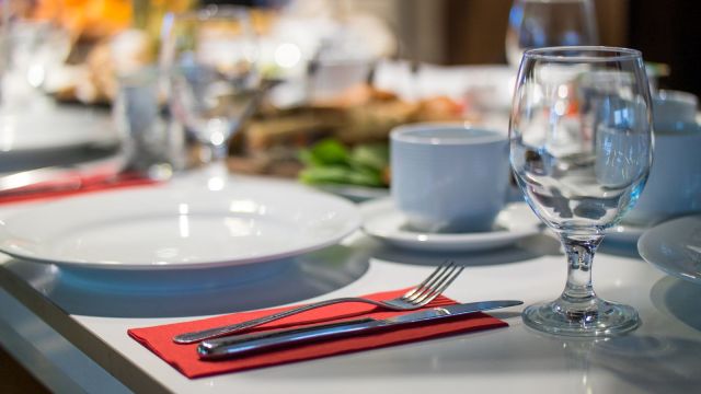 Masă de restaurant aranjată elegant, cu farfurii albe, pahar de vin și tacâmuri pe un șervet roșu