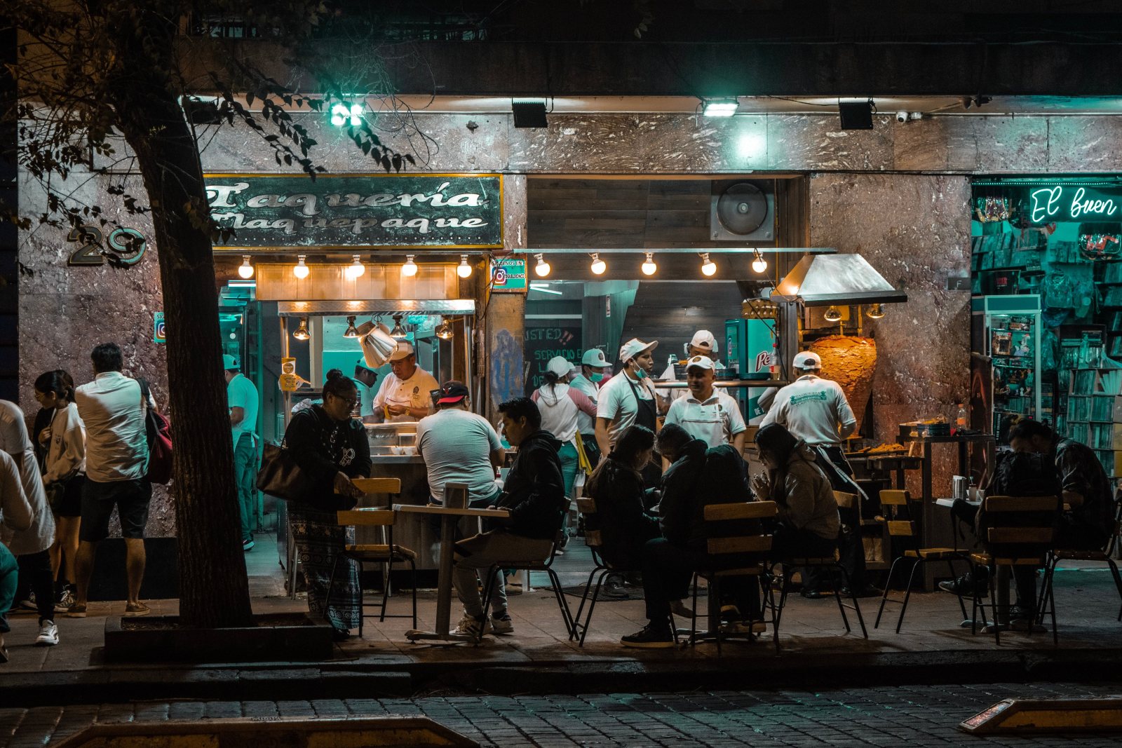 Oameni la mesele unui restaurant în aer liber, seara, într-o stradă din oraș luminată